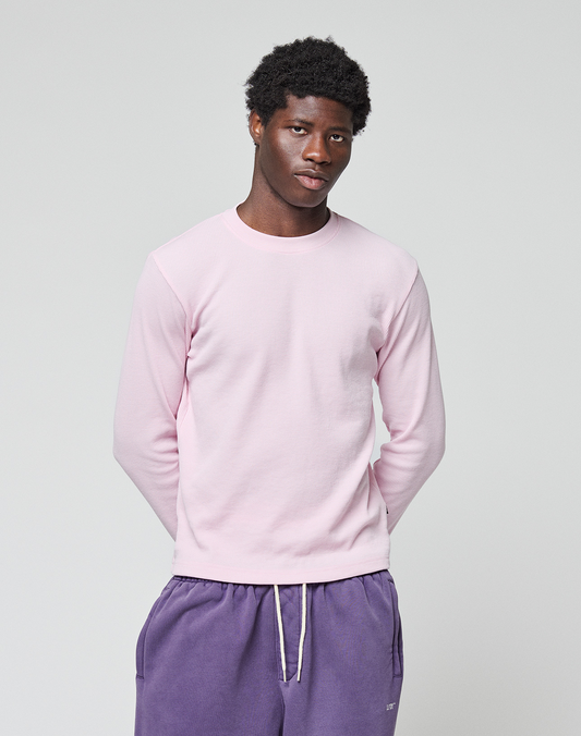 A young man with short curly hair wears the LIVE FAST DIE YOUNG Basic Waffle Longsleeve in light pink and purple drawstring cotton pants, standing against a plain light gray background with his hands behind his back.