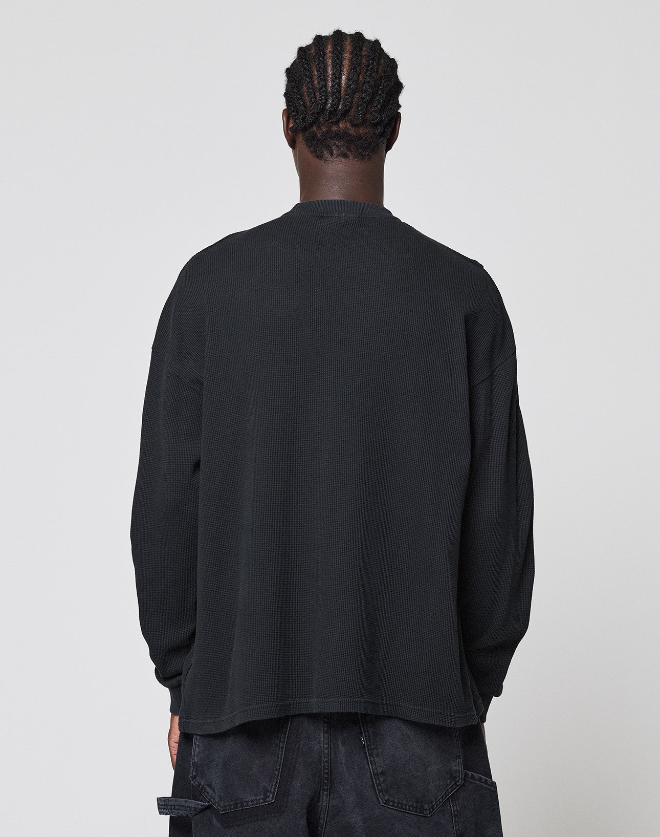 A person with short, braided hair faces away from the camera, wearing a LIVE FAST DIE YOUNG Blank Waffle Longsleeve in black and black pants with Ripp-Bündchen, set against a plain light gray background.