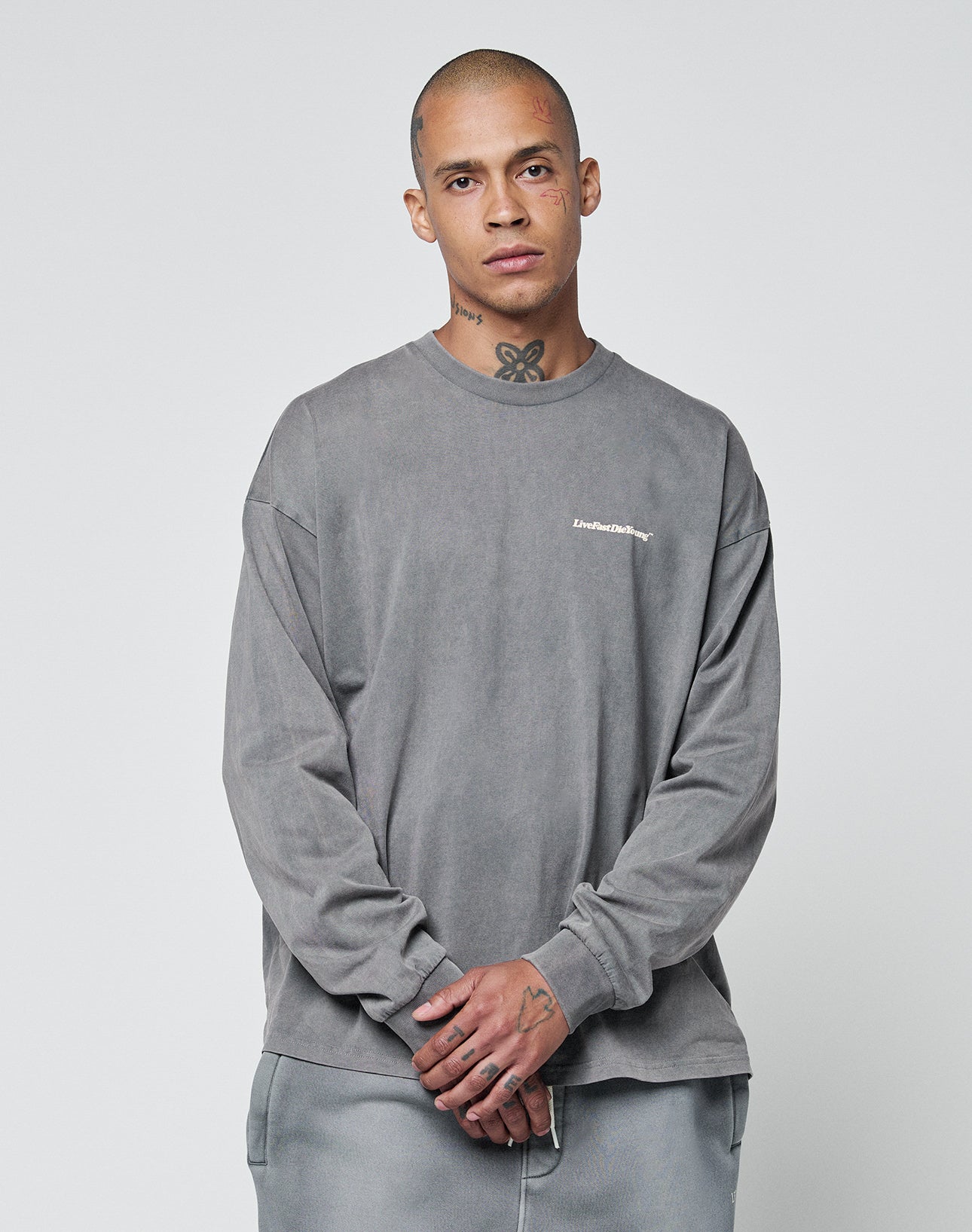 A shaved-headed person with face and neck tattoos wears a loose-fitting, 100% Baumwolle Basic Longsleeve and matching pants with subtle logo-print by LIVE FAST DIE YOUNG, standing against a plain light background, hands clasped in front.