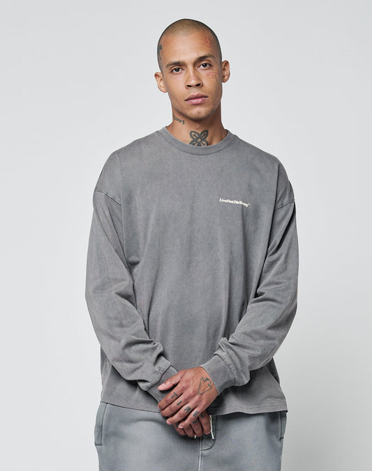 A shaved-headed person with face and neck tattoos wears a loose-fitting, 100% Baumwolle Basic Longsleeve and matching pants with subtle logo-print by LIVE FAST DIE YOUNG, standing against a plain light background, hands clasped in front.