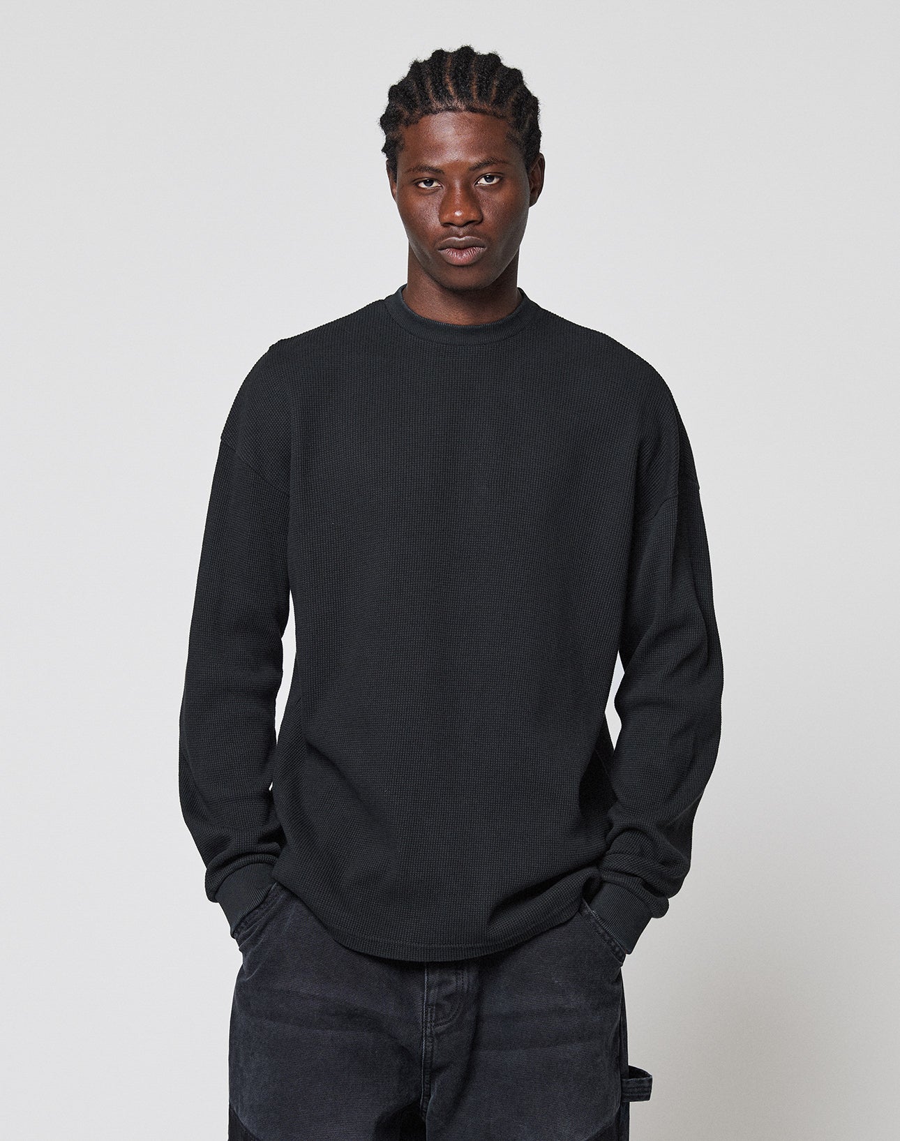 A man with braided hair wears the LIVE FAST DIE YOUNG Blank Waffle Longsleeve aus 100% Baumwolle with Ripp-Bündchen, paired with dark pants, hands in his pockets, standing before a light gray background and looking at the camera.