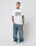A tattooed man faces the camera in a white LIVE FAST DIE YOUNG LFDY Sixers Tee (Regular Fit, 100% Baumwolle), loose blue jeans, and white sneakers against a gray background. Hands relaxed by his sides. Waschhinweise included.