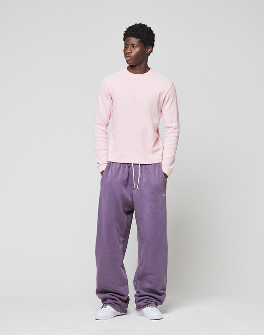 A person stands against a plain light background, wearing the LIVE FAST DIE YOUNG Basic Waffle Longsleeve in light pink, loose purple sweatpants with ribbed cuffs and white drawstring, and white sneakers, looking slightly to the side.