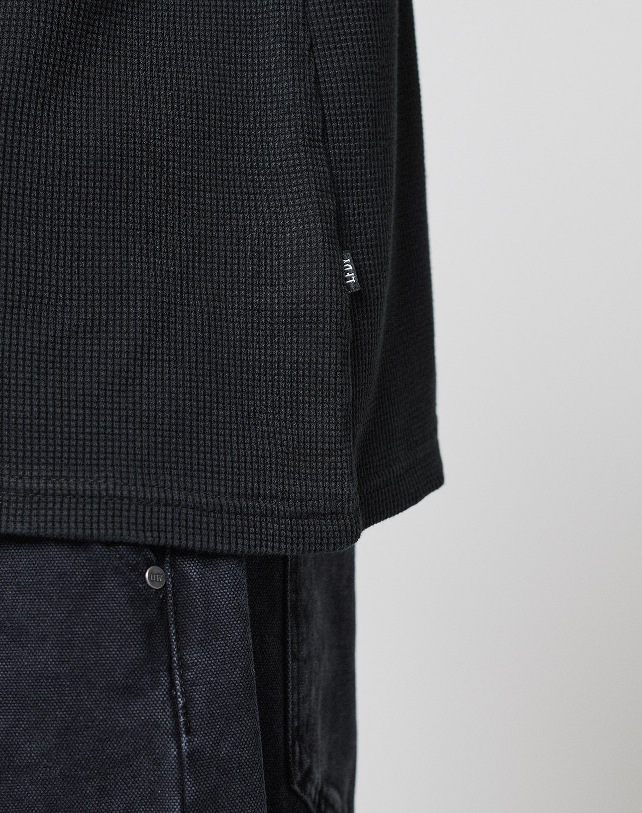 Close-up of the LIVE FAST DIE YOUNG Blank Waffle Longsleeve in black with textured fabric, Ripp-Bündchen, and small side tag, styled with dark denim pants against a plain light backdrop.