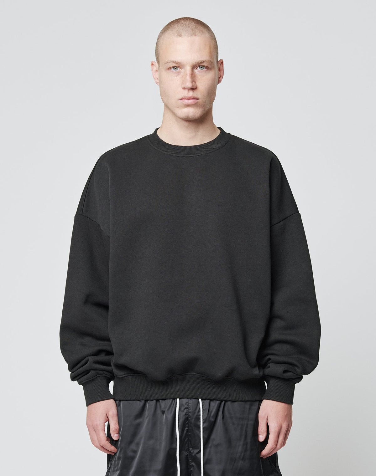 A person with a buzz cut is wearing the LIVE FAST DIE YOUNG Basic Blank Sweater, an Oversize Fit black sweatshirt made from soft Baumwolle and Polyester, paired with black shorts, standing against a plain light gray background. | model