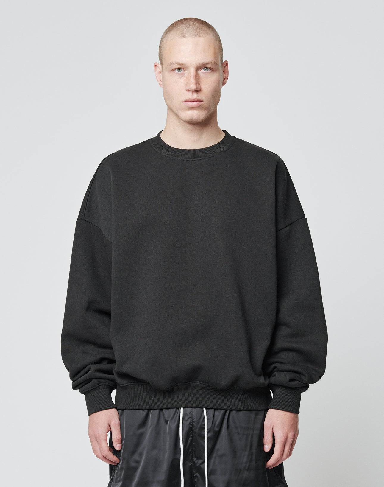 A person with a buzz cut is wearing the LIVE FAST DIE YOUNG Basic Blank Sweater, an Oversize Fit black sweatshirt made from soft Baumwolle and Polyester, paired with black shorts, standing against a plain light gray background. | model