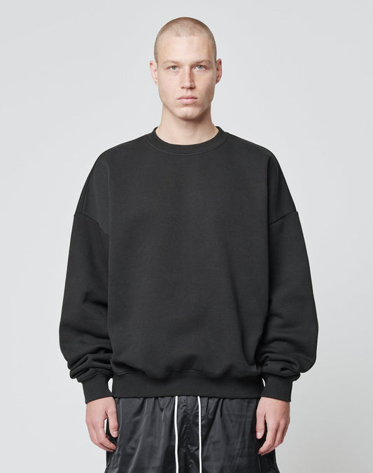 A person with a buzz cut is wearing the LIVE FAST DIE YOUNG Basic Blank Sweater, an Oversize Fit black sweatshirt made from soft Baumwolle and Polyester, paired with black shorts, standing against a plain light gray background. | model