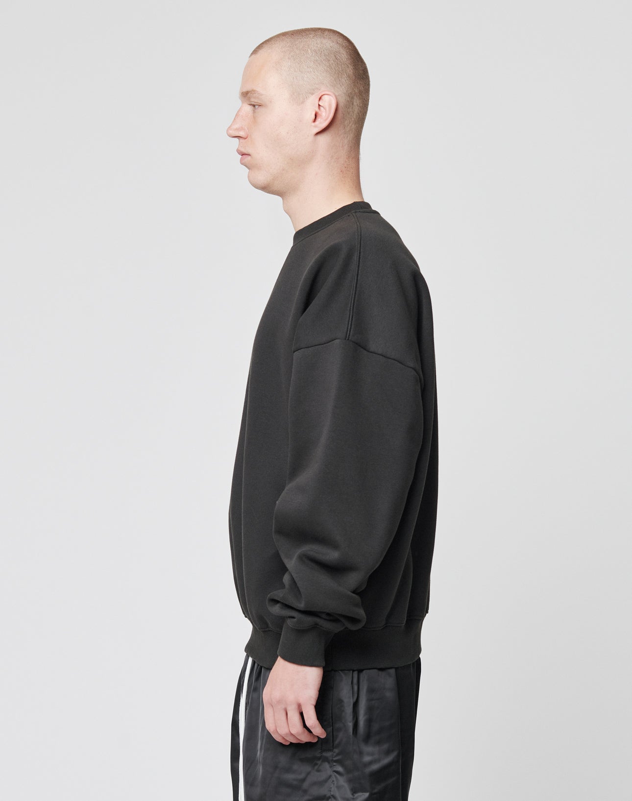 A person with a shaved head stands in profile against a plain light gray background, wearing an oversize fit Basic Blank Sweater and black pants by LIVE FAST DIE YOUNG, both crafted from soft Baumwolle.
