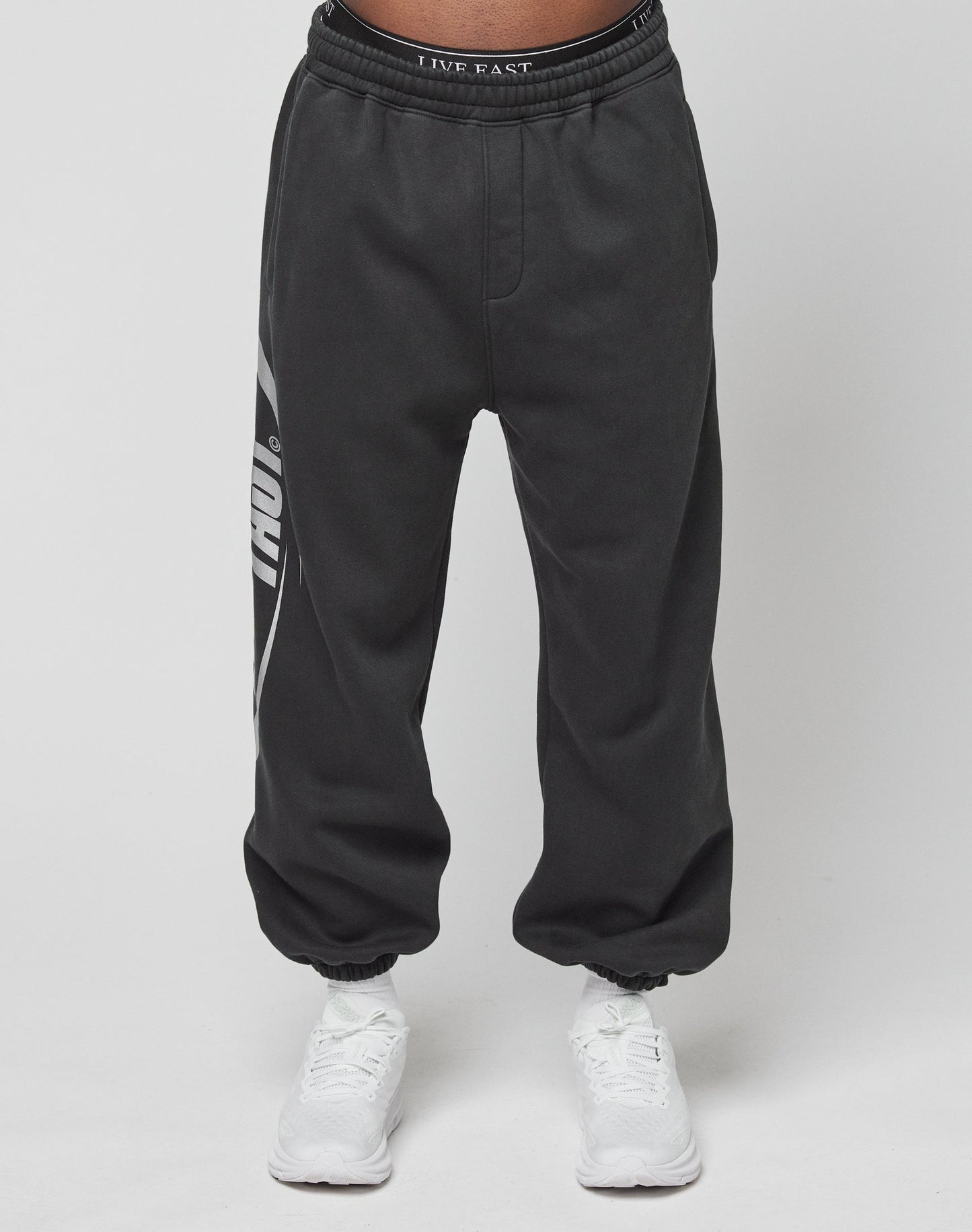 A person wears LIVE FAST DIE YOUNG’s LFDY Sphere Jogger, black with a white left-leg graphic and an Elastikbund waistband reading LIVE FAST, paired with white sneakers against a plain white background.