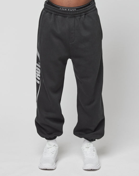 A person wears LIVE FAST DIE YOUNG’s LFDY Sphere Jogger, black with a white left-leg graphic and an Elastikbund waistband reading LIVE FAST, paired with white sneakers against a plain white background.