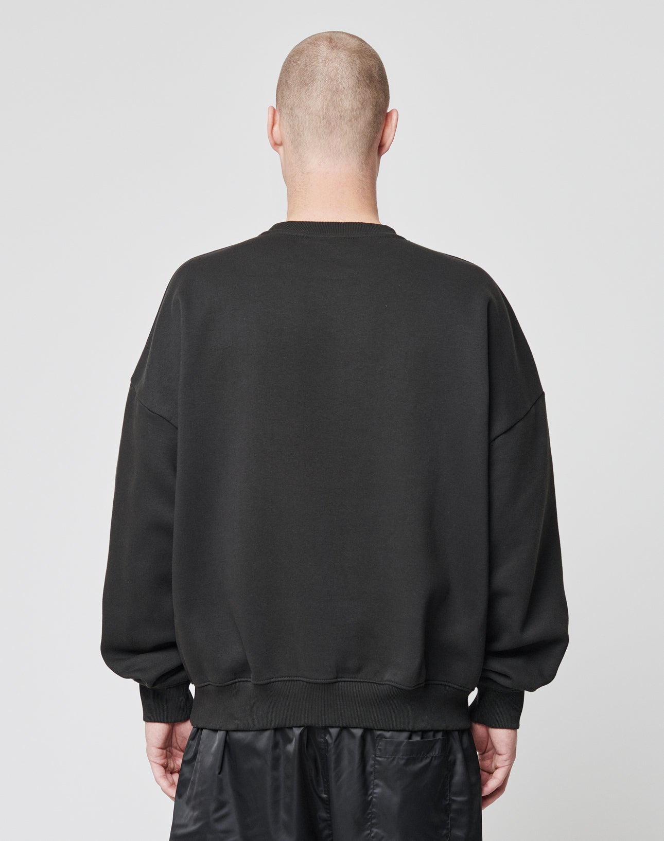 A person with a shaved head faces away from the camera, wearing a LIVE FAST DIE YOUNG Basic Blank Sweater in black and black shorts, set against a plain light background.