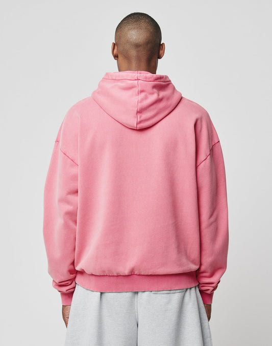 A person faces away from the camera, wearing a LIVE FAST DIE YOUNG Ripped College Hooded in pink with the hood down and oversized light gray pants, set against a plain light gray background.