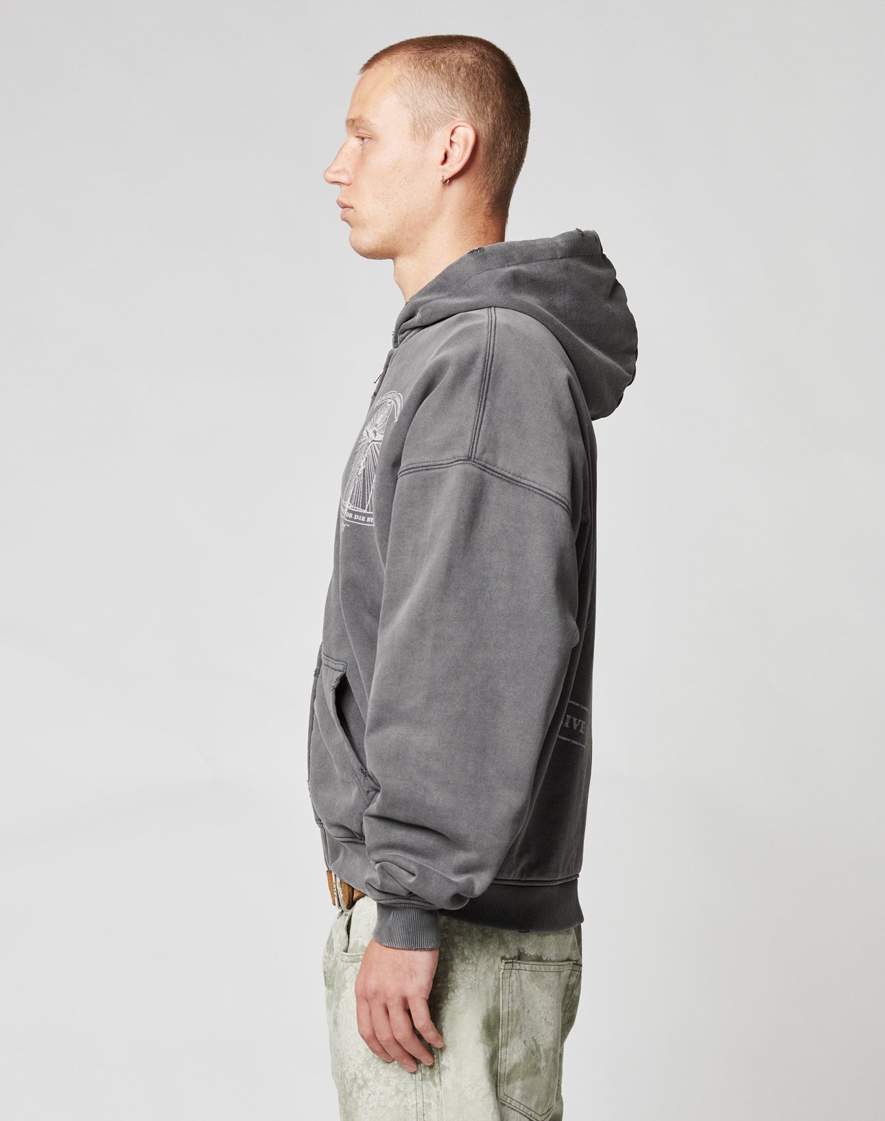 A man with short hair stands in profile, wearing a faded gray LIVE FAST DIE YOUNG LFDY Cross Hooded Zip and light-colored pants against a plain, light-colored background.