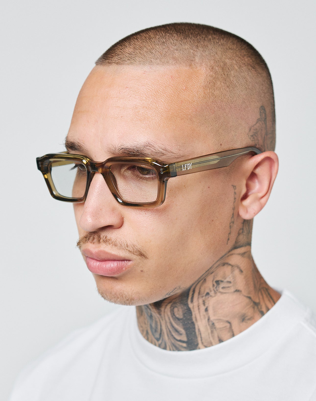 A person with close-cropped hair and tattoos wears LIVE FAST DIE YOUNG LFDY x Chimi Shades 03 sunglasses and a white shirt, looking slightly to the side against a plain background.