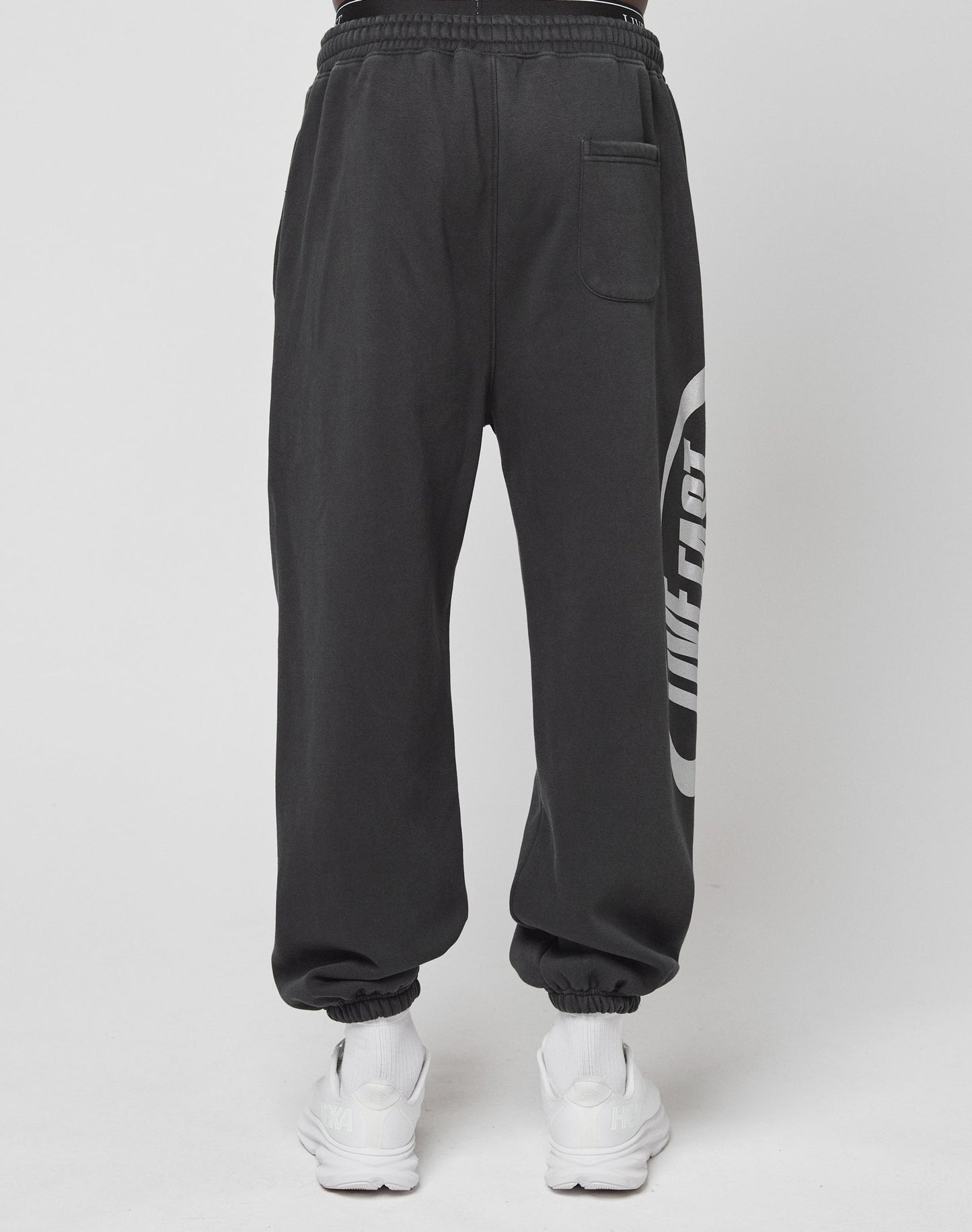 A person wears black LFDY Sphere Jogger pants by LIVE FAST DIE YOUNG, featuring a back pocket, Oversize Fit, and distinctive side logo, paired with white sneakers, standing against a plain white background.