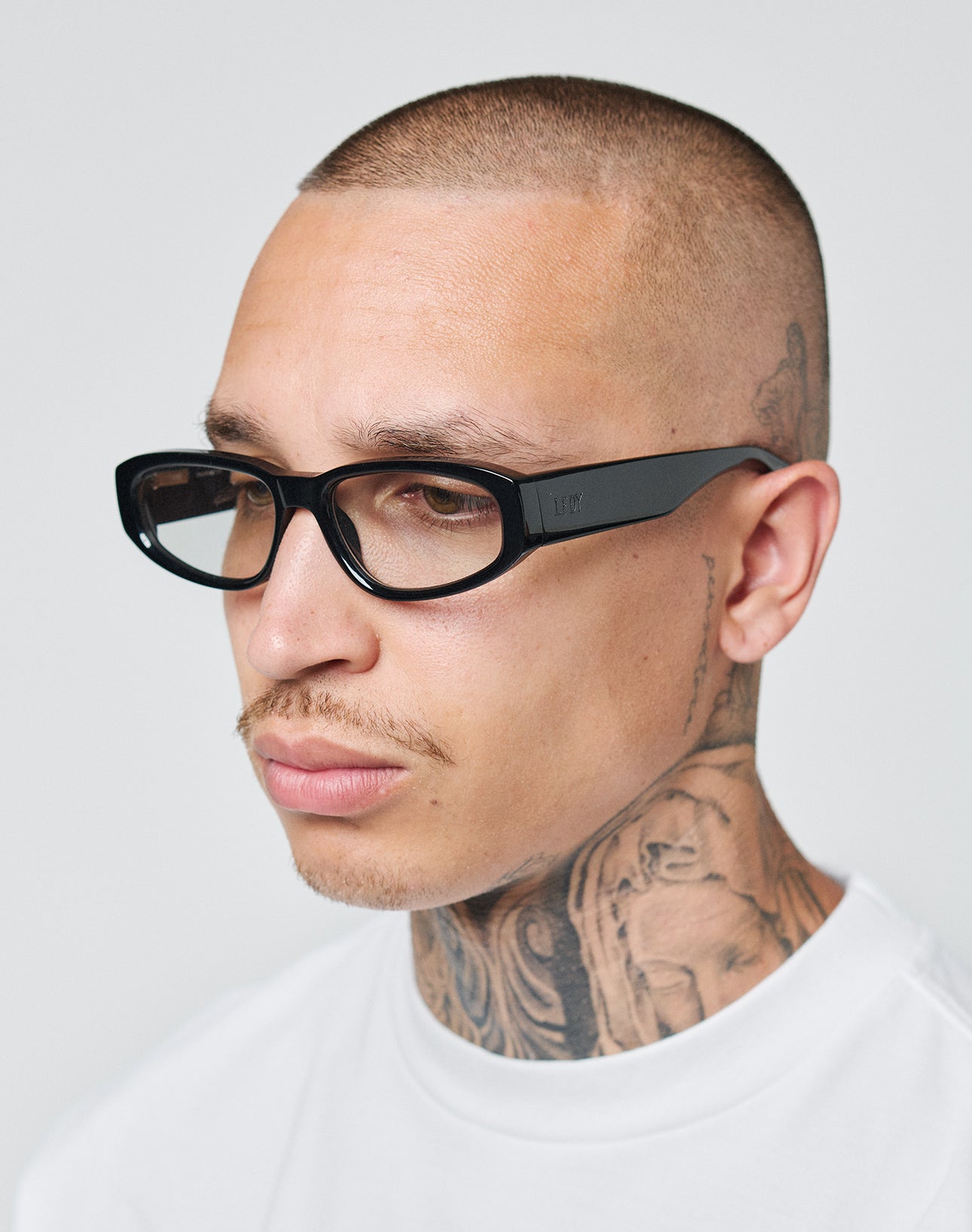 A person with a shaved head, neck and scalp tattoos, wearing a white shirt and LFDY x Chimi Shades 09 by LIVE FAST DIE YOUNG, looks slightly to the side against a plain background.
