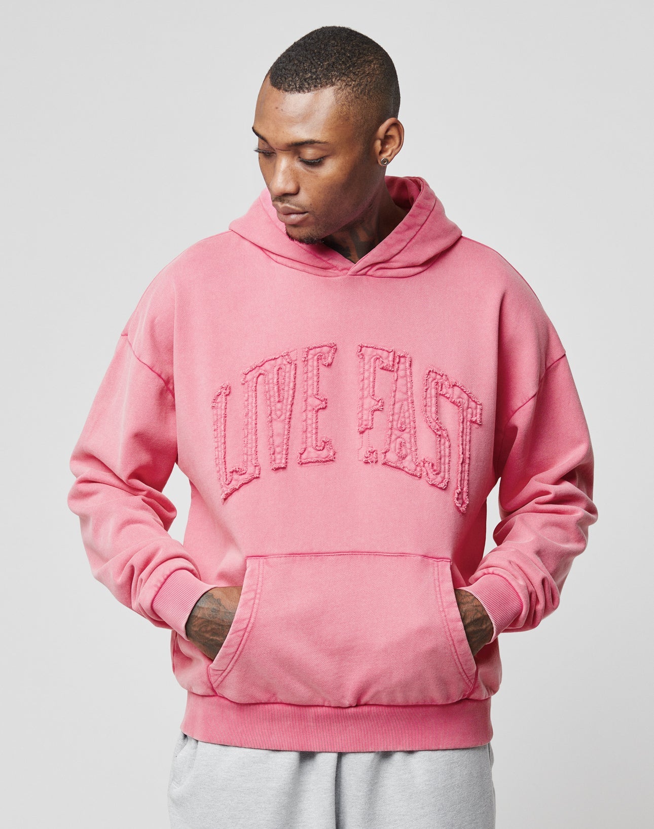 A person wears the LIVE FAST DIE YOUNG Ripped College Hooded in pink, featuring distressed LIVE FAST text and an oversize fit. Made from 100% Baumwolle, it’s styled with light gray pants and hands-in-pocket, gazing downward.