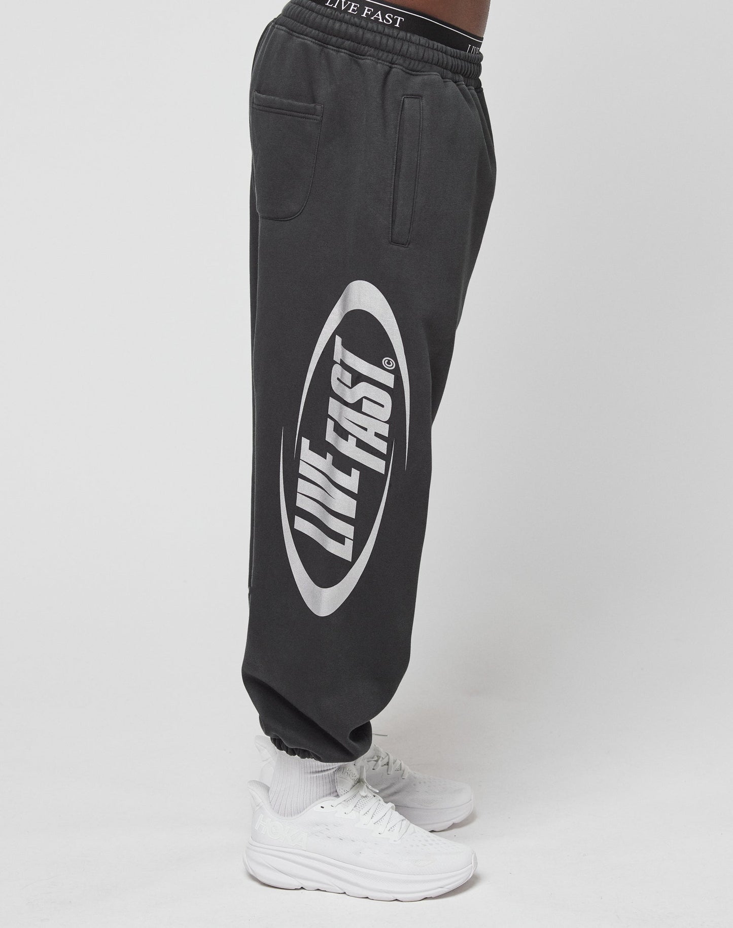 A person wearing LFDY Sphere Jogger black sweatpants by LIVE FAST DIE YOUNG with an Elastikbund, oversize fit, and a large white “LIVE FAST” logo on the side, paired with white sneakers against a plain white background.