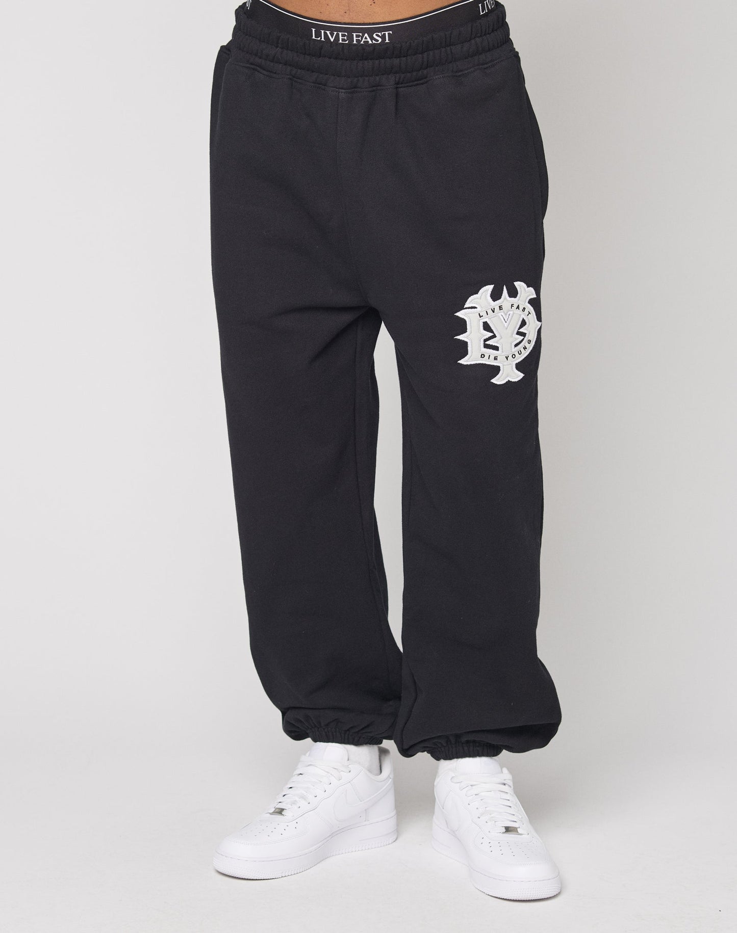 A person wears the DY Loose Jogger by LIVE FAST DIE YOUNG—schwarze Baumwollhose mit weißem Graphic am linken Oberschenkel, Elastikbund mit "LIVE FAST" Schriftzug—kombiniert mit weißen Sneakern vor weißem Hintergrund.