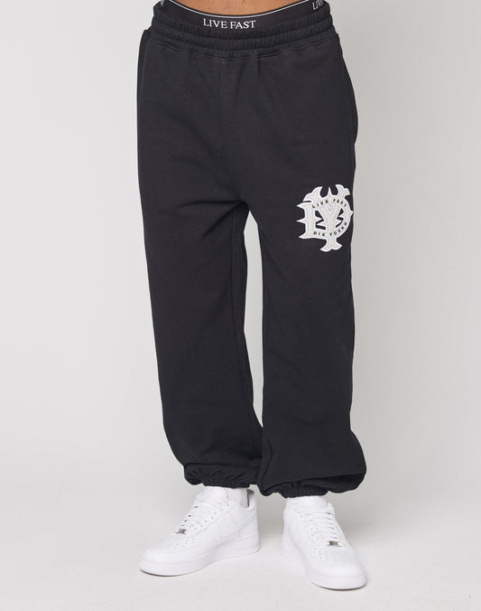 A person wears LIVE FAST DIE YOUNG DY Jogger, an oversize fit black sweatpant with a white left-thigh graphic and elastikbund, pairing it with white sneakers and black LIVE FAST waistband underwear. The background is plain and light.