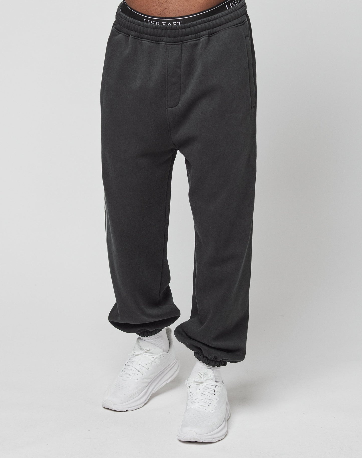 Wearing the LFDY Sphere Jogger by LIVE FAST DIE YOUNG—black cotton sweatpants with an oversize fit, elastic waistband featuring LIVE FAST, and cuffs—paired with white sneakers against a plain white background.