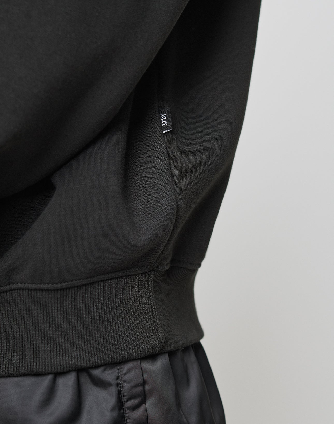 Close-up of a person wearing the LIVE FAST DIE YOUNG Basic Blank Sweater in black, featuring a ribbed waistband made from soft Baumwolle and a small black tag with white text on the side seam.