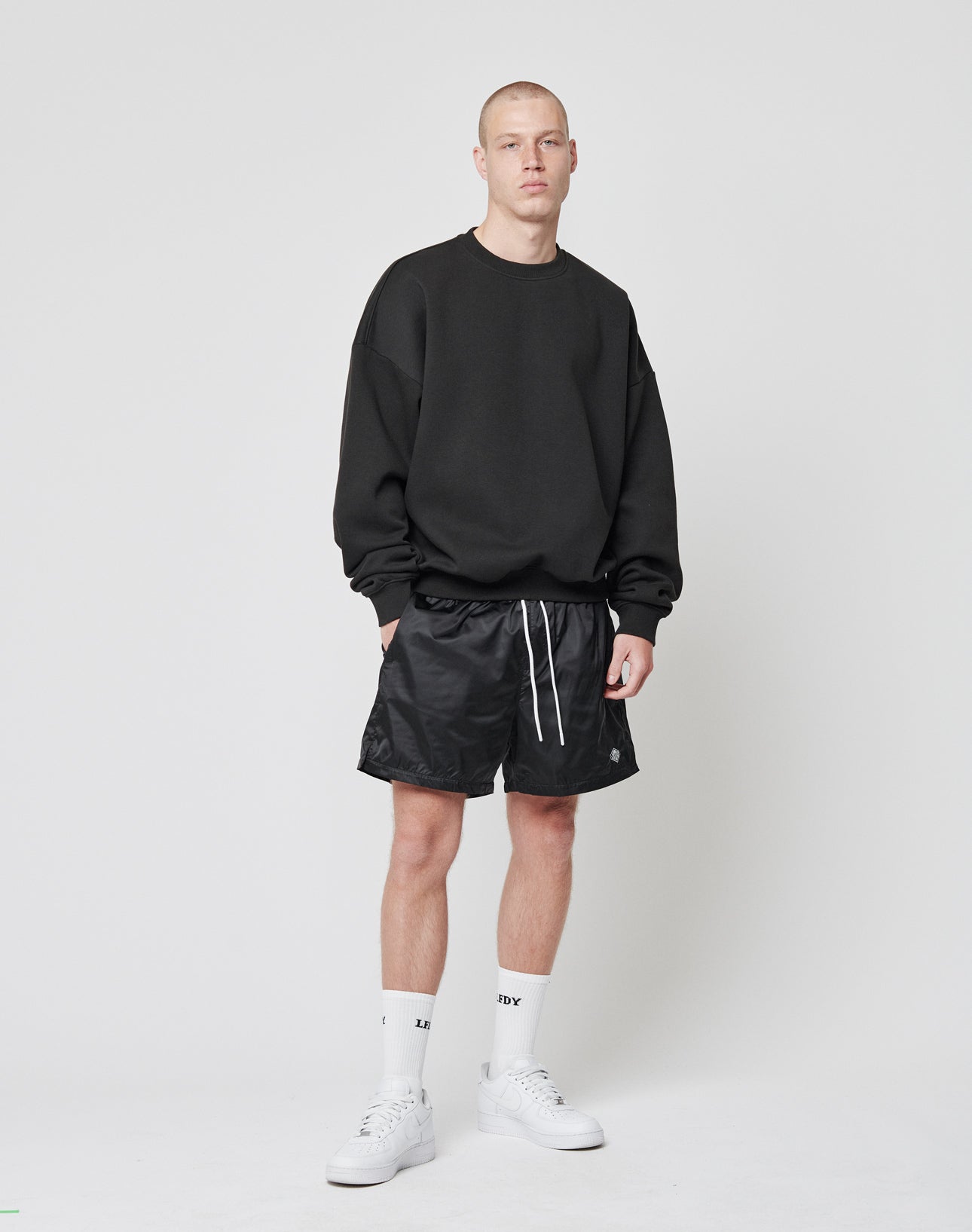 A person with a neutral expression stands against a white backdrop wearing the LIVE FAST DIE YOUNG Basic Blank Sweater in an oversize fit, paired with black athletic shorts, white crew socks, and white sneakers.