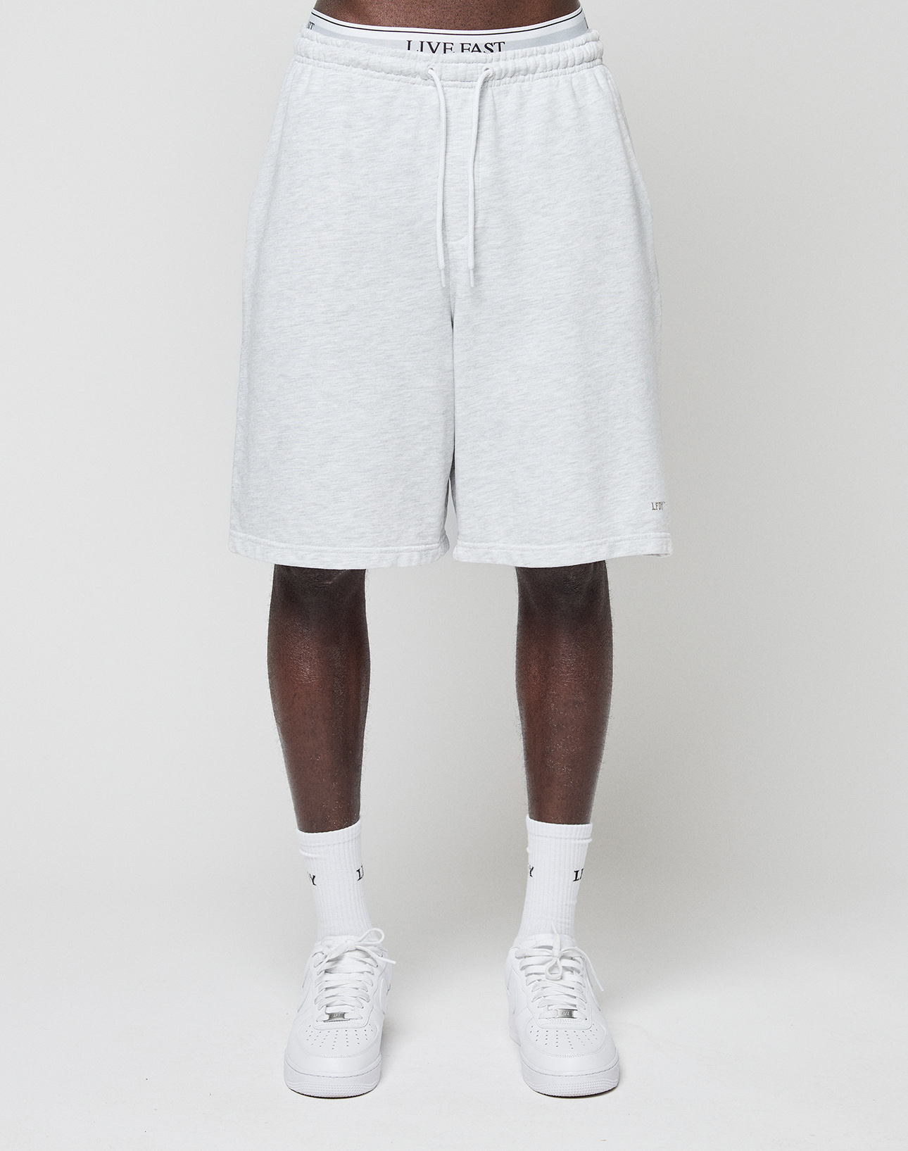 A person wears LIVE FAST DIE YOUNG Basic Shorts in light gray with elastic waistband and "LIVE FAST" logo print, along with white crew socks and sneakers, standing against a plain white background. The upper body is not visible.