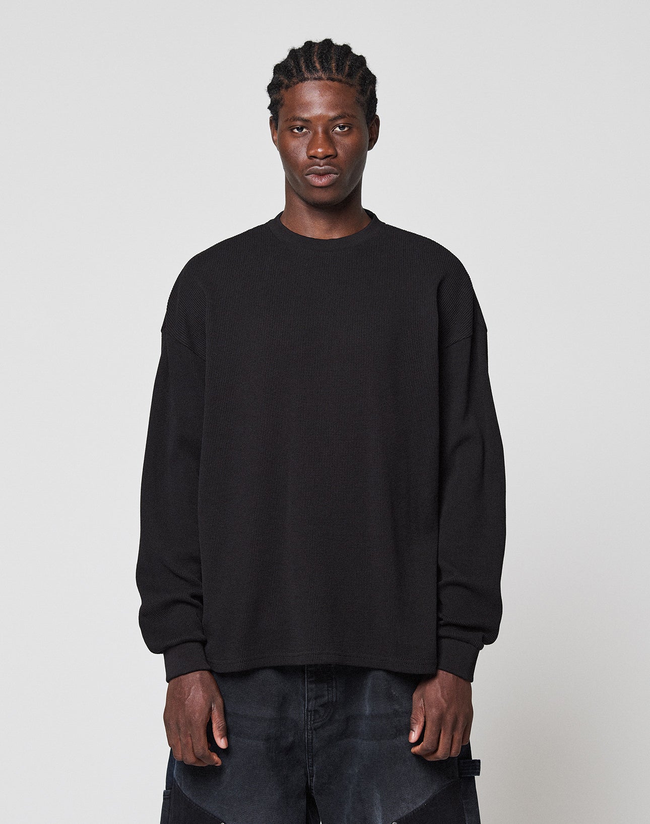 A person with short braided hair and a neutral expression stands against a plain light gray background, wearing the LIVE FAST DIE YOUNG Blank Waffle Longsleeve in black with Ripp-Bündchen and dark pants. | model