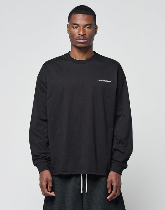A man faces forward in a LIVE FAST DIE YOUNG Basic Longsleeve—black, loose-fit, with subtle logo text on the left chest—paired with black shorts. The oversize style looks relaxed against a plain light gray background. | model