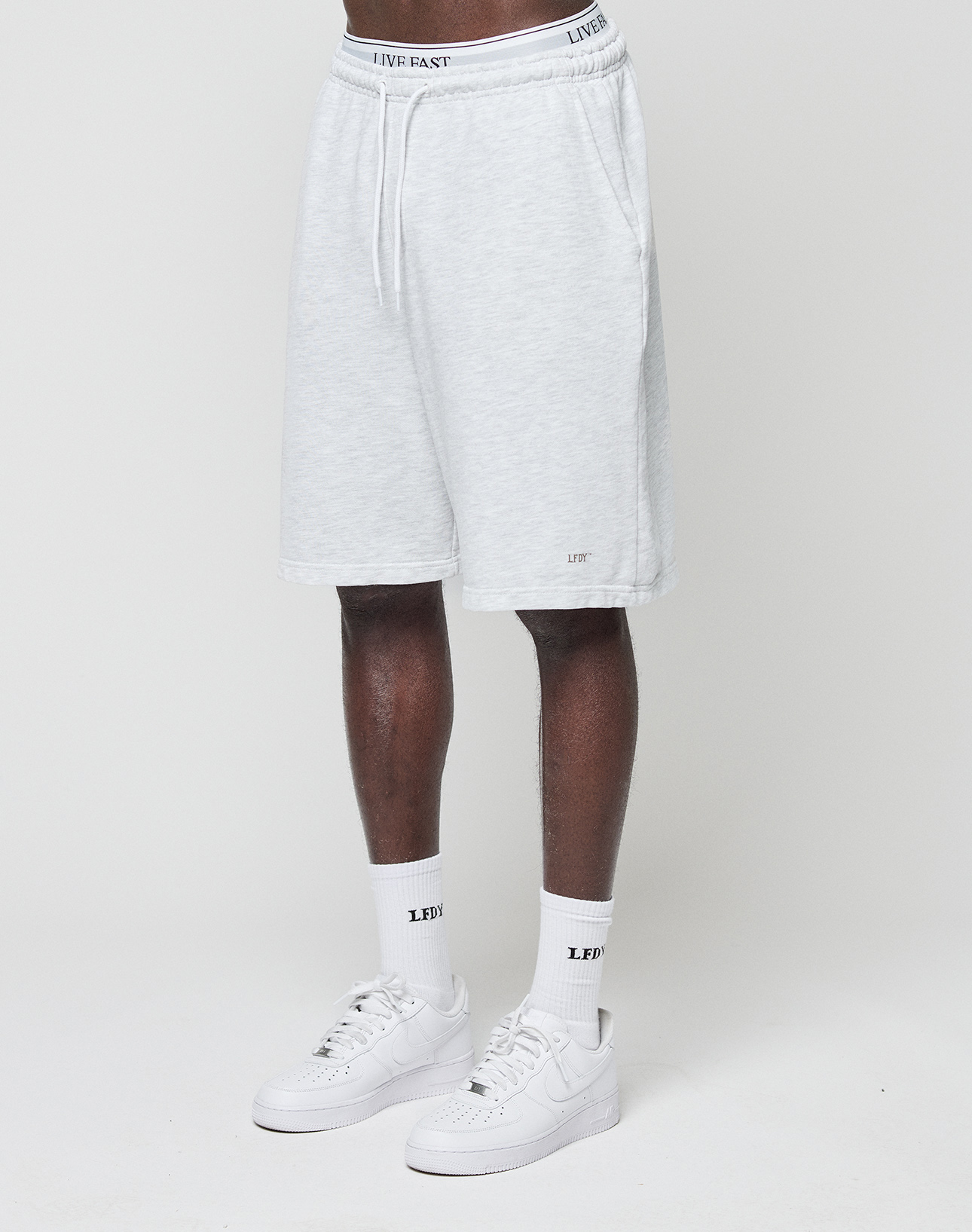 A person wears LIVE FAST DIE YOUNG Basic Shorts in regular fit with elastic waistband and logo print, paired with white LFDY socks and white Nike sneakers, standing against a plain white background.