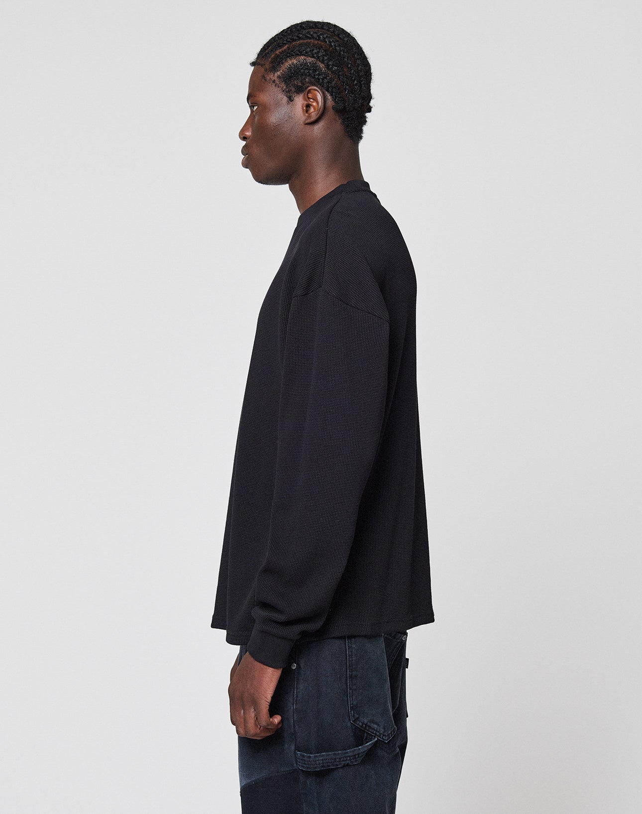 A person with braided hair stands in profile against a plain, light background, wearing an oversize fit LIVE FAST DIE YOUNG Blank Waffle Longsleeve in black with Ripp-Bündchen and dark pants.