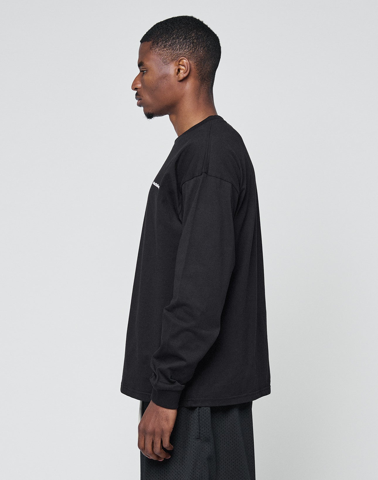 A man in profile faces left, dressed in the LIVE FAST DIE YOUNG Basic Longsleeve—100% Baumwolle black shirt—and oversize-fit black textured pants, against a plain light gray background.
