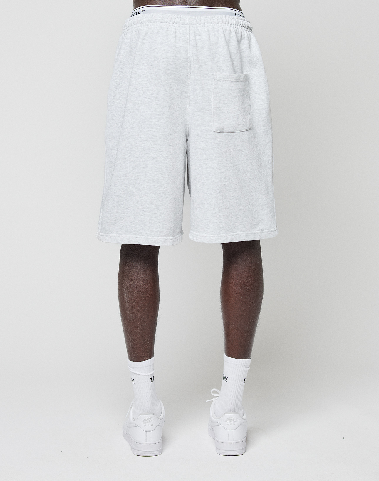 A person faces away, wearing LIVE FAST DIE YOUNG Basic Shorts in light gray with elastic waistband and back pocket, paired with white socks and white sneakers, set against a plain light background.