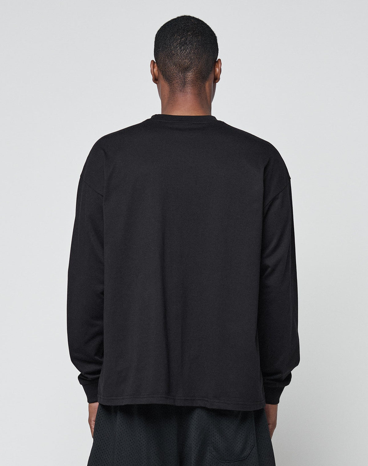 A person faces away from the camera, wearing LIVE FAST DIE YOUNGs Basic Longsleeve—an oversized black 100% Baumwolle shirt—and black shorts, all set against a light gray background.