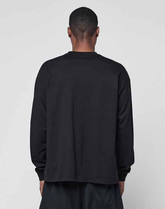 A person faces away from the camera, wearing LIVE FAST DIE YOUNGs Basic Longsleeve—an oversized black 100% Baumwolle shirt—and black shorts, all set against a light gray background.