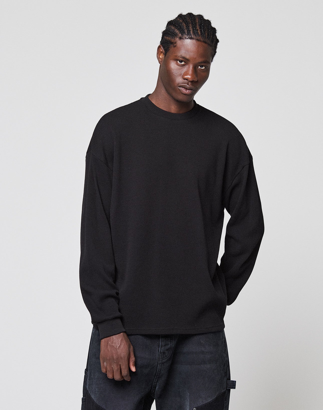 A young man with braided hair stands against a plain background, wearing a LIVE FAST DIE YOUNG Blank Waffle Longsleeve in an oversize fit and black jeans, his expression neutral.