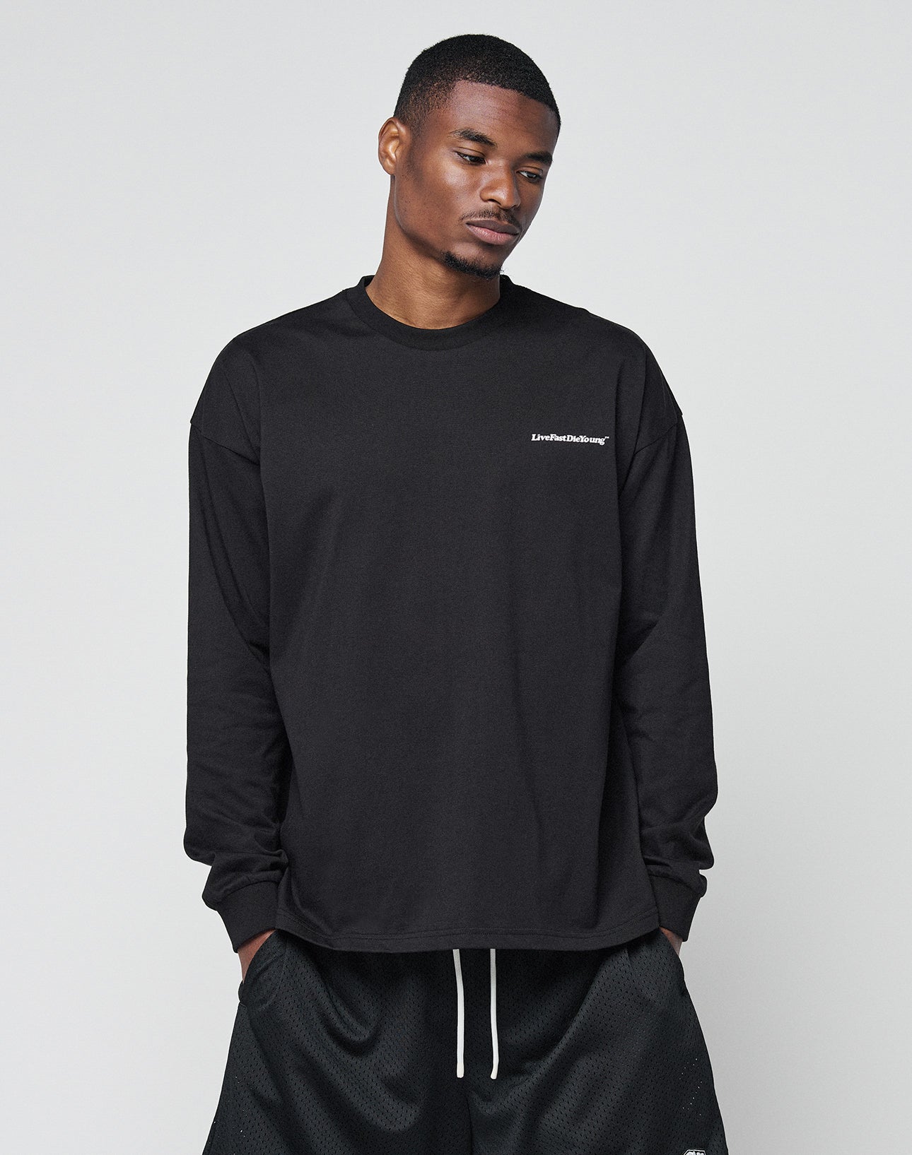 A man stands against a light gray background, looking downward with hands in his pockets, wearing the LIVE FAST DIE YOUNG Basic Longsleeve—an oversized black shirt with white chest logo—and black shorts.