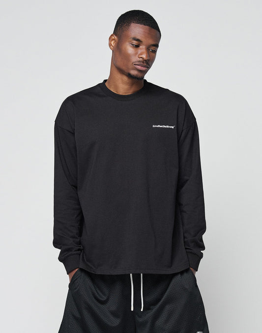 A man stands against a light gray background, looking downward with hands in his pockets, wearing the LIVE FAST DIE YOUNG Basic Longsleeve—an oversized black shirt with white chest logo—and black shorts.