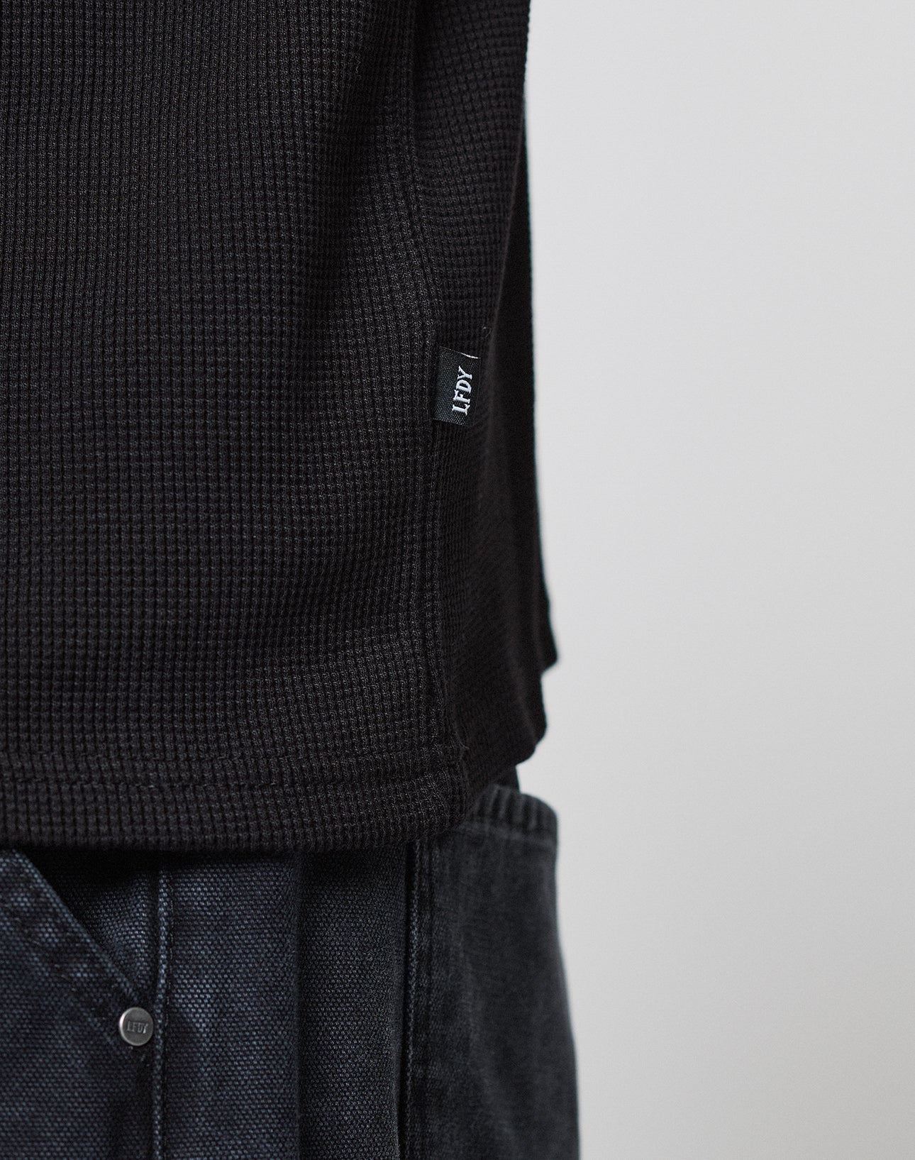 Close-up of a person wearing the LIVE FAST DIE YOUNG Blank Waffle Longsleeve, a black textured shirt with Ripp-Bündchen and a HEMP fabric tag near the hem, paired with dark denim pants against a light plain background.