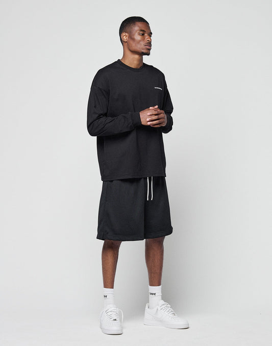 A man stands against a plain light gray background wearing a black LIVE FAST DIE YOUNG Basic Longsleeve, black knee-length shorts, white socks, and white sneakers. He is facing slightly right with his hands clasped together.