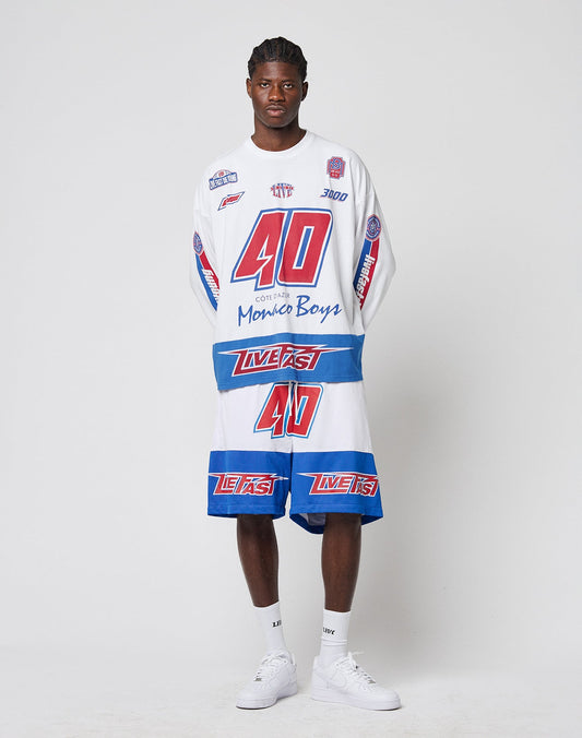A man stands against a plain background wearing LIVE FAST DIE YOUNG’s LFDY Racing Mesh Shorts and a matching racing-style shirt, both featuring large prints, bold red, blue, and white graphics, numbers, and text. He pairs them with white sneakers and socks.