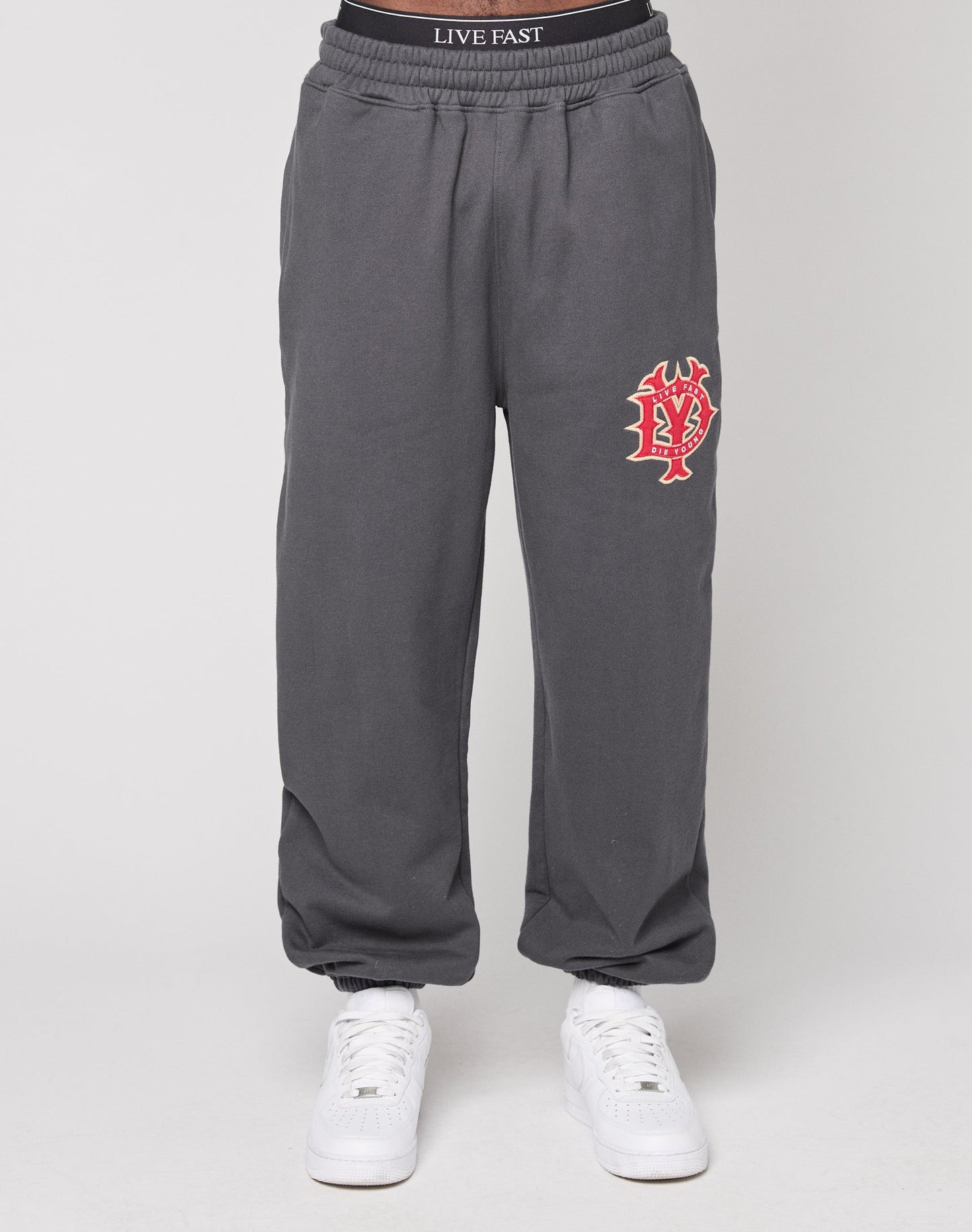 A person wears charcoal gray DY Loose Jogger by LIVE FAST DIE YOUNG with a red and white graphic logo on the left thigh, white sneakers, and an elastic waistband that says LIVE FAST, posed against a plain background.