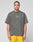 A man with braided hair wears the LIVE FAST DIE YOUNG Live Fast Wave Tee, an oversized, faded gray T-shirt with a small LIVE FAST reflective front print, paired with bright yellow shorts, standing against a light gray background.