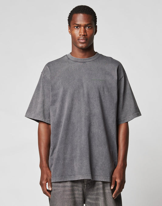 A man faces forward against a plain light gray background, wearing the LIVE FAST DIE YOUNG LFDY Hardwear Tee in Oversize Fit, a loose-fitting faded black T-shirt, paired with matching dark jeans.