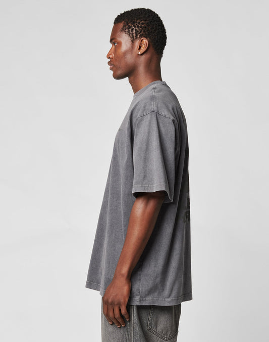 A young man stands in profile against a plain background, wearing the LIVE FAST DIE YOUNG LFDY Hardwear Tee with an oversize fit and gray jeans. His arms are by his sides as he looks ahead.