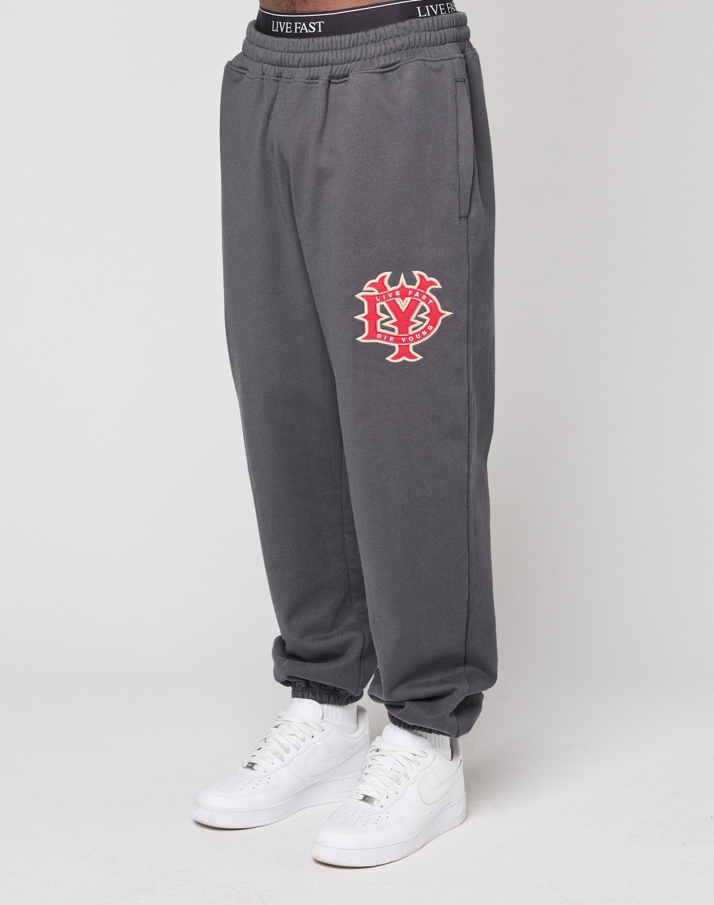 A person shows only their lower body, hands in pockets, wearing LIVE FAST DIE YOUNG's gray DY Loose Jogger with a red and white graphic on the left thigh and white sneakers. The sweatpants have an Elastikbund and are set against a plain background.