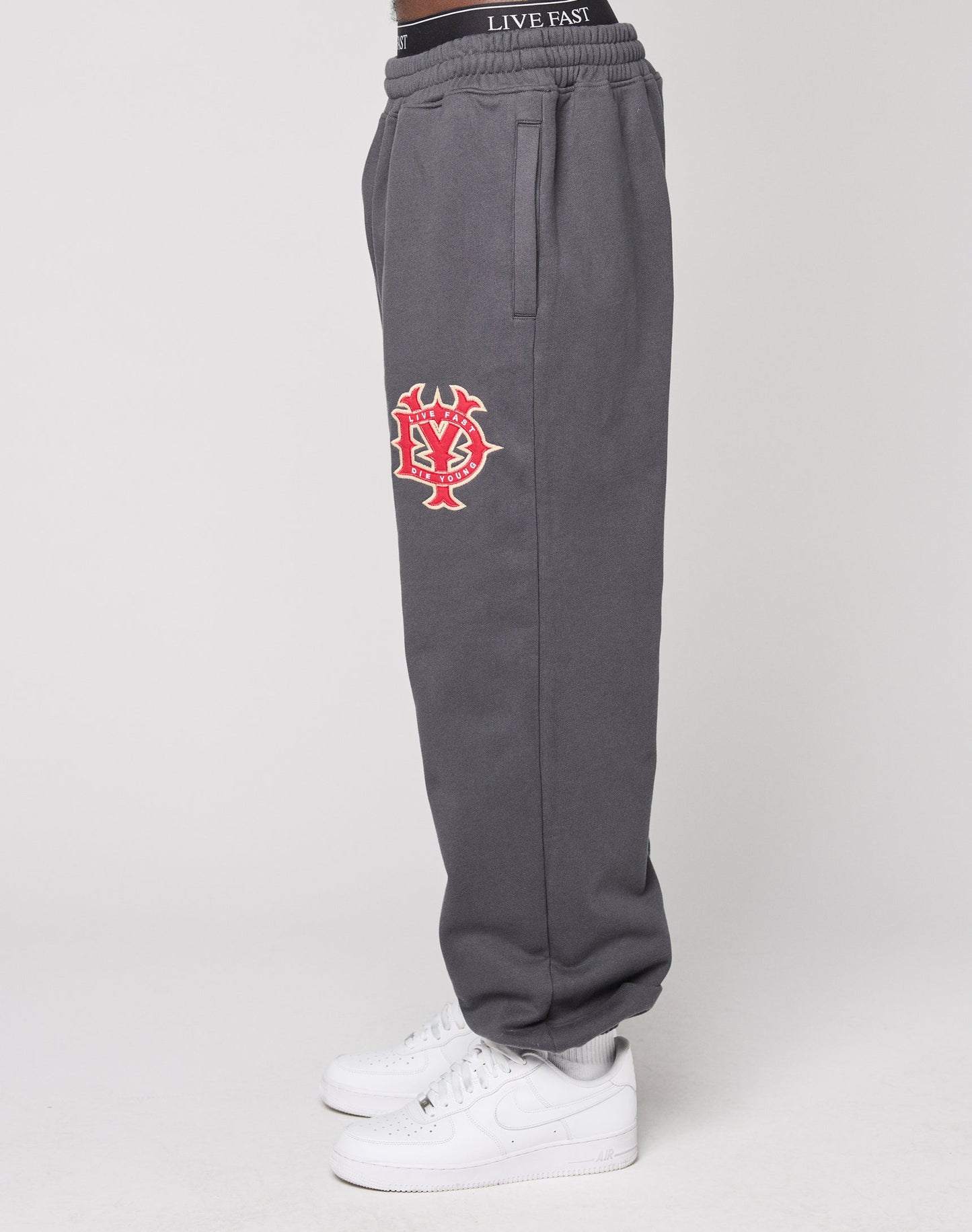 A person wears gray DY Jogger Sweatpants by LIVE FAST DIE YOUNG with elastic waistband and red, white embroidered logo on the thigh, paired with white socks and white Nike sneakers, standing in profile against a neutral background.