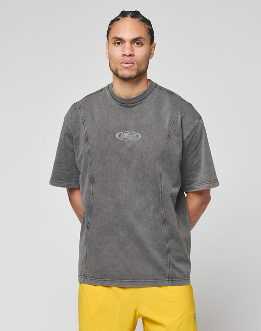 A man with braided hair wears the LIVE FAST DIE YOUNG Live Fast Wave Tee— a loose-fitting, faded gray T-shirt with reflective front print— and bright yellow shorts, standing against a light gray background with his hands behind his back.