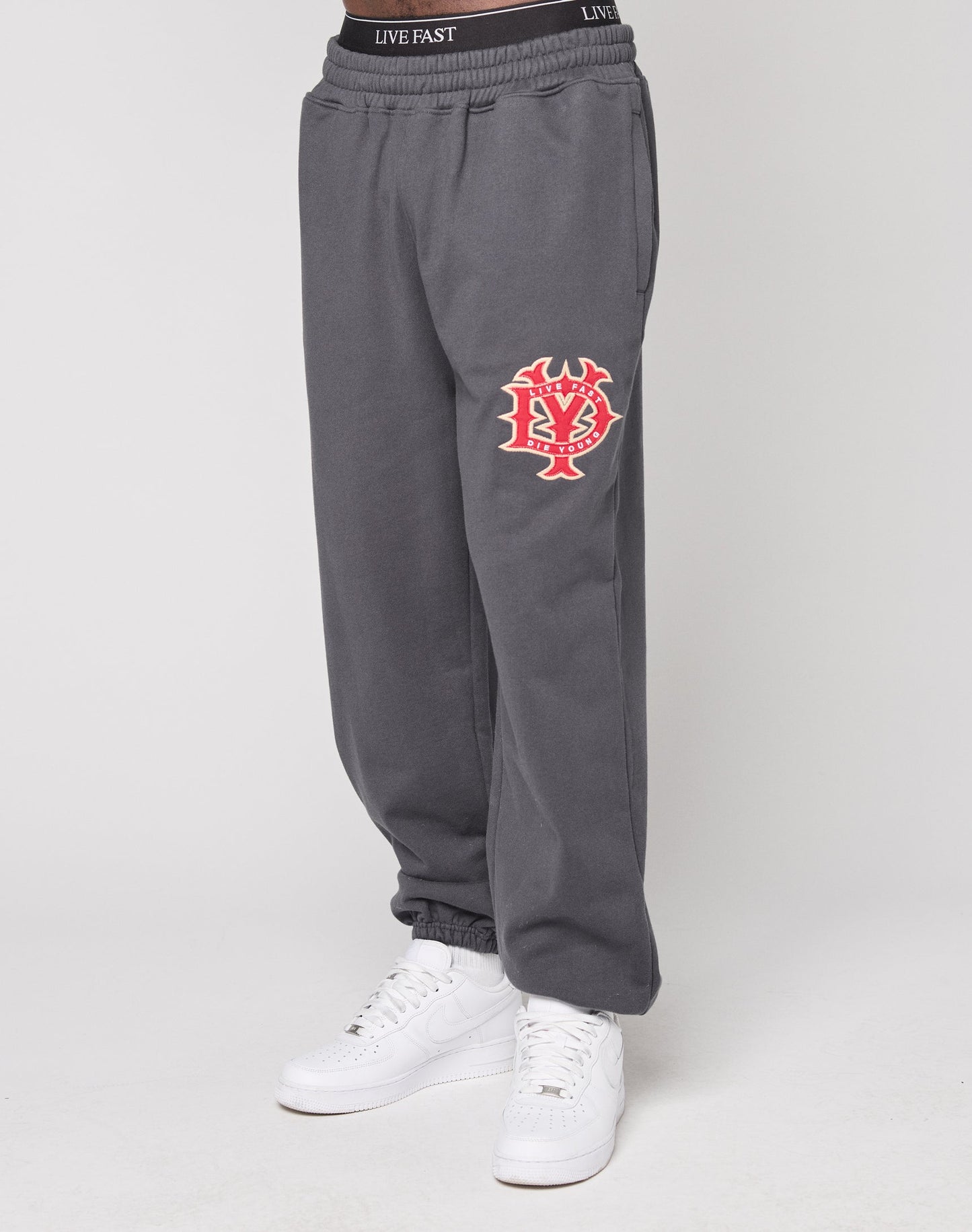 A person wearing the DY Loose Jogger from LIVE FAST DIE YOUNG—dark gray cotton sweatpants with an elastic waistband, orange and white graphic patch on the left thigh, and "LIVE FAST" printed in white—paired with white sneakers, against a plain light background.